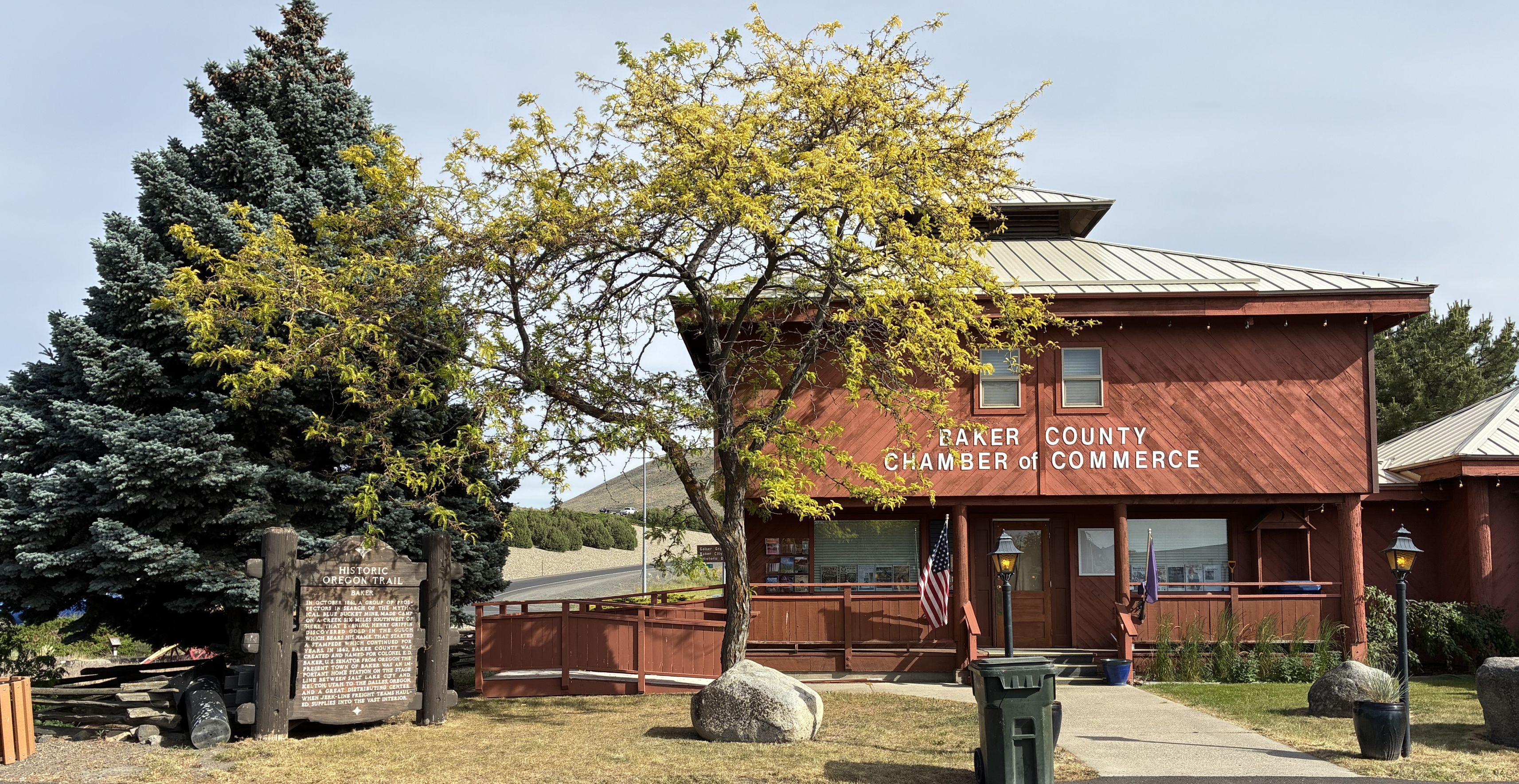 Image of old wooden sign in front of pine tree, Baker Chamber office to right.