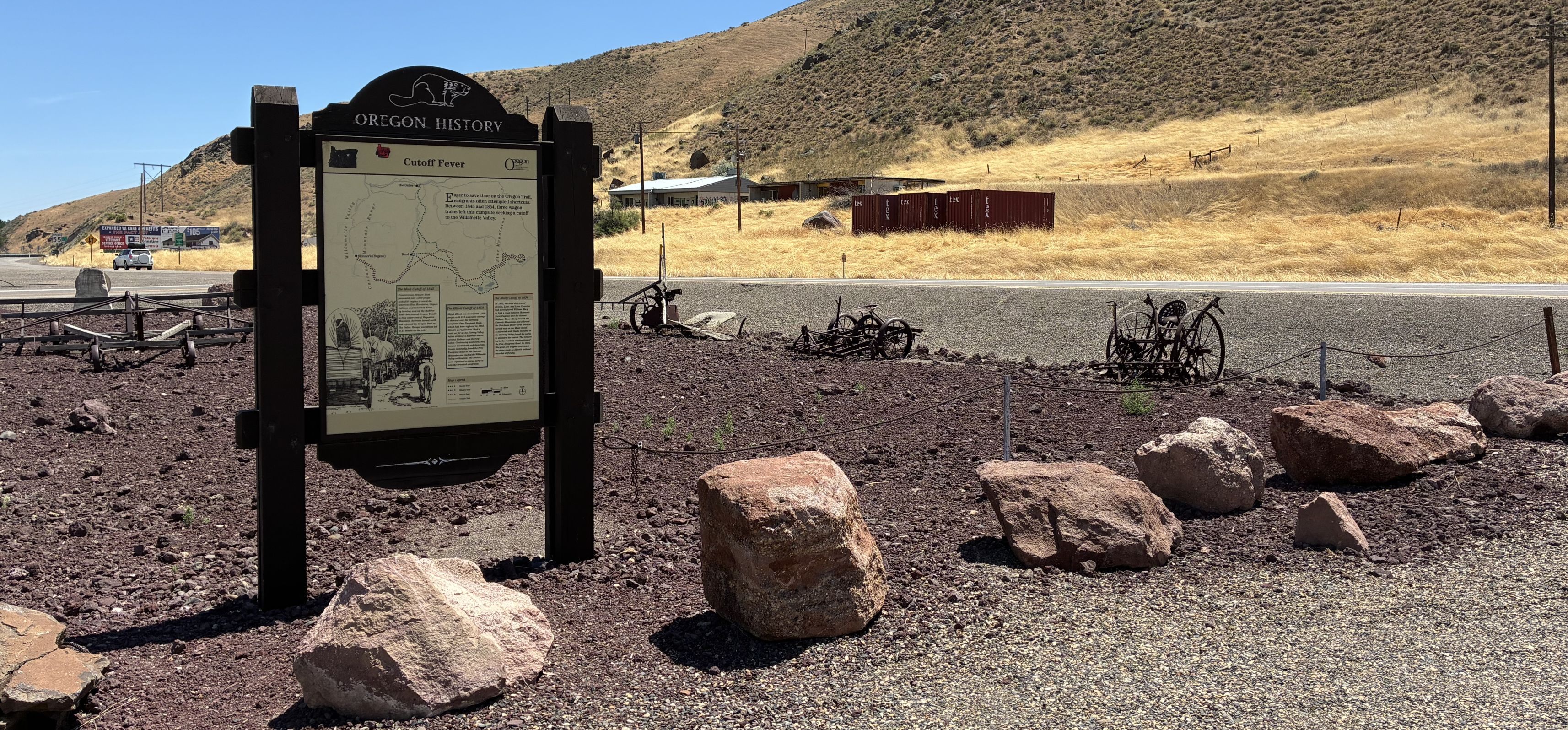 Wooden sign in open rocky area, boulders in foreground and dry grass covered hill in background.