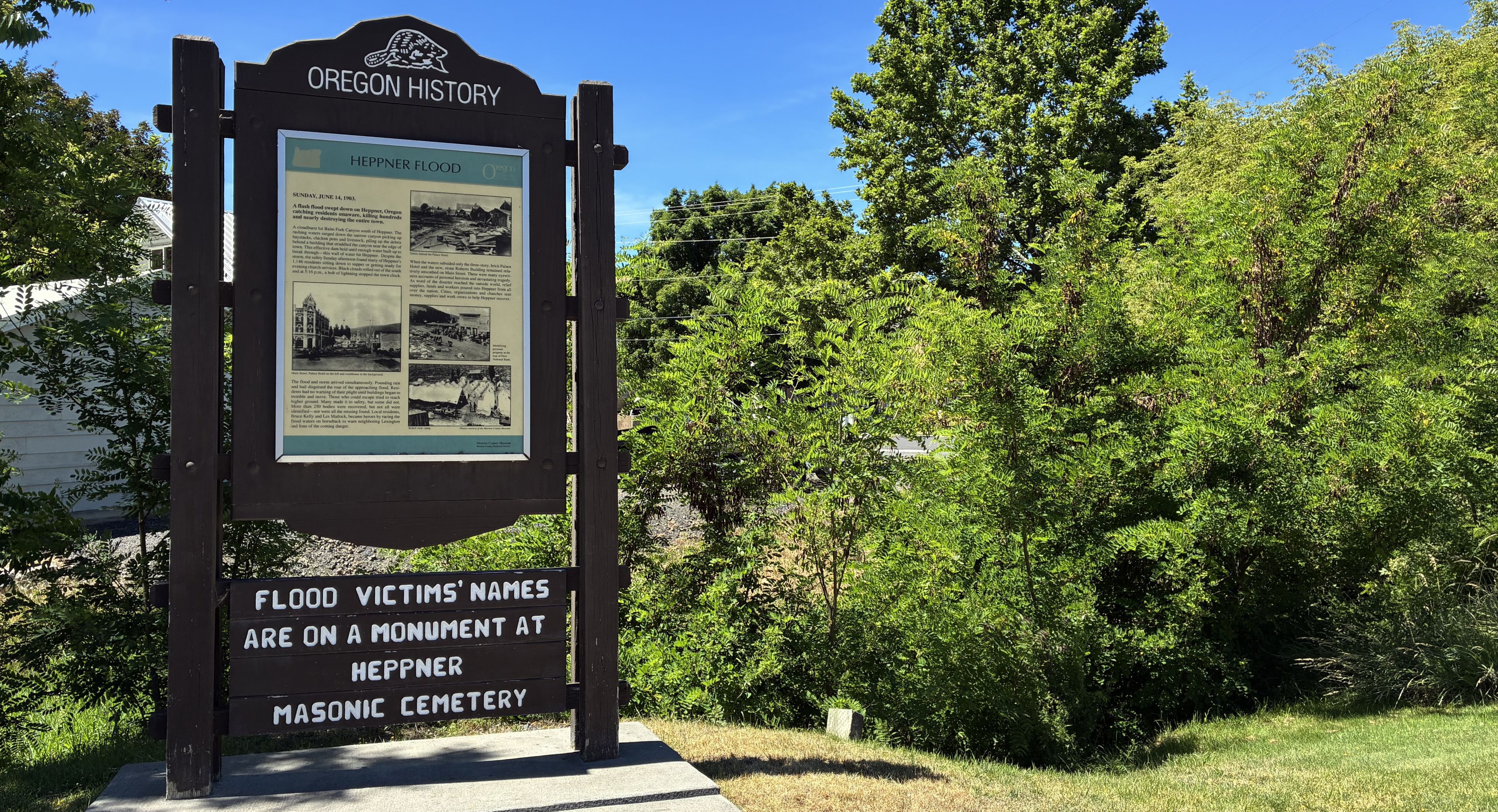 Wooden sign next to green trees and sidewalk in city park.