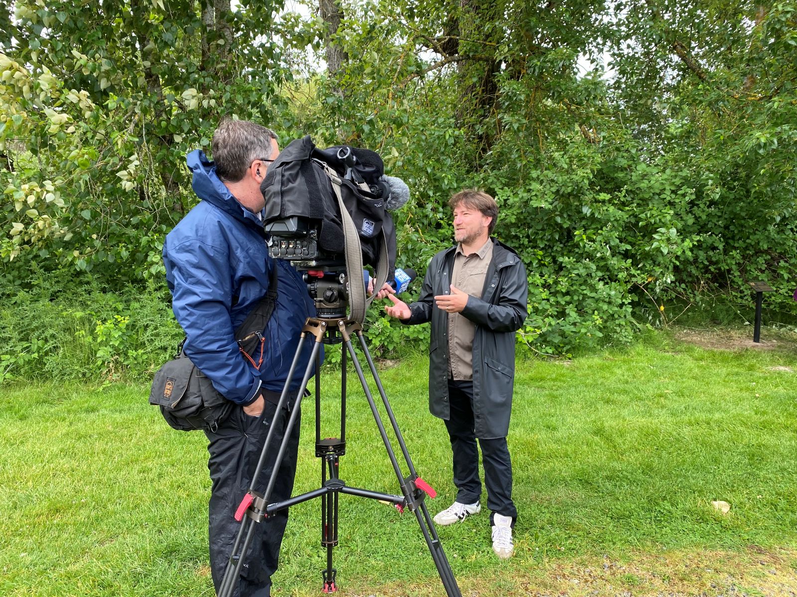 Reporter in blue coat standing behind video camera interviewing man in black rain coat. Green trees in background.